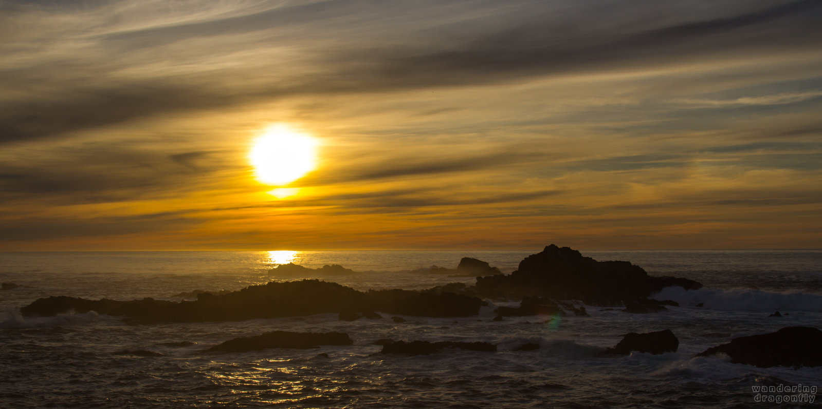 Blurred descending sun -- cloud, ocean, rock, sun, sunset, water, wave